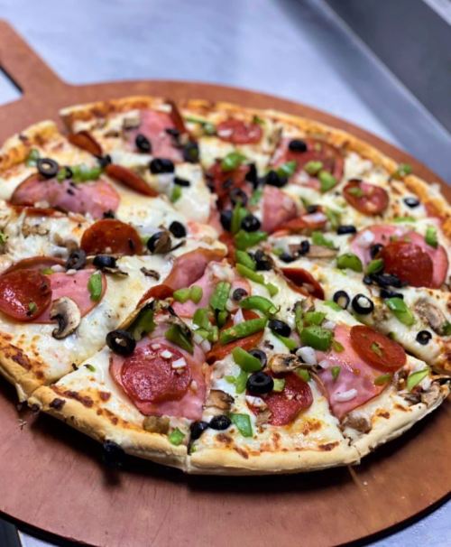 A freshly baked pizza topped with pepperoni, ham, mushrooms, olives, and green peppers on a wooden pizza board.