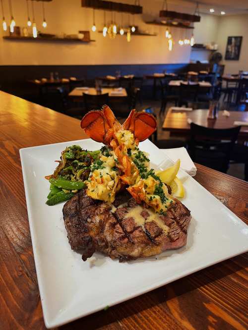 A gourmet plate featuring a grilled steak, lobster tail, sautéed greens, and lemon wedges, set in a cozy restaurant.