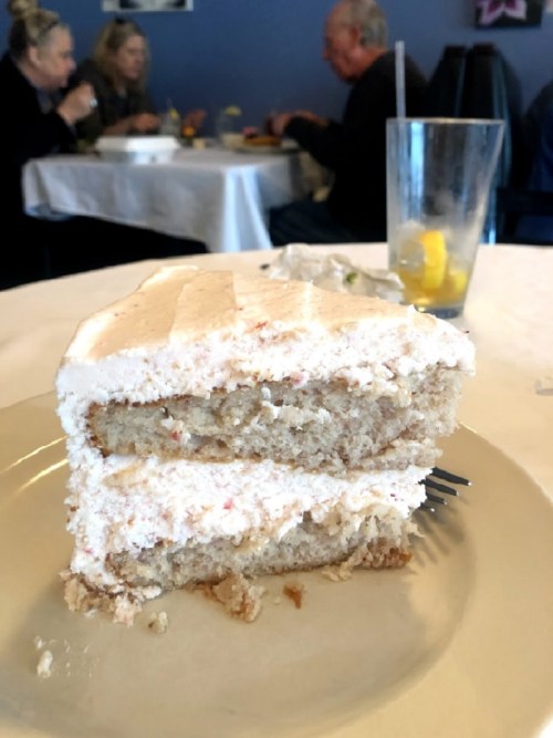 A slice of pink frosted cake on a plate, with a blurred restaurant background and people dining.