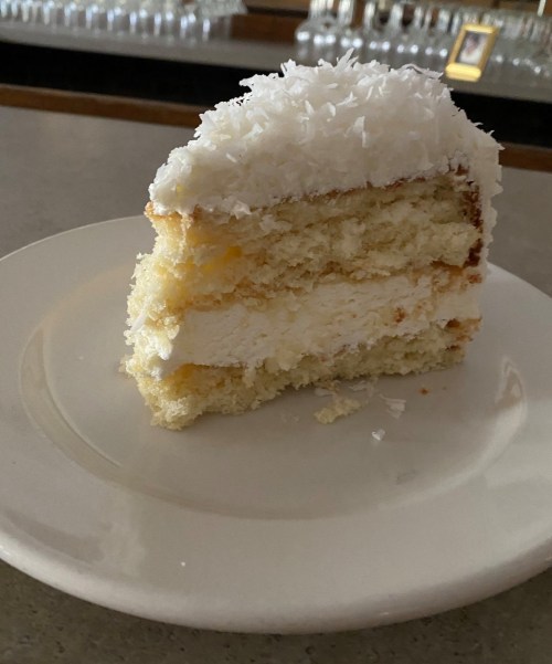A slice of coconut cake with layers of cream and a fluffy coconut topping on a white plate.