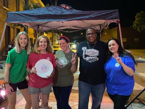 Five people stand together under a tent at night, smiling and holding a plate, with colorful shirts and a festive atmosphere.