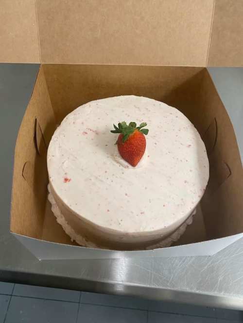 A round cake with a smooth white frosting and a single strawberry on top, placed inside a brown cardboard box.