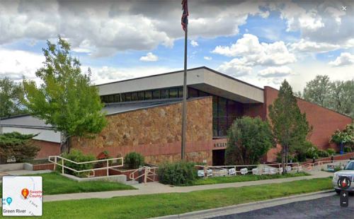 A modern building with a stone facade, surrounded by greenery and a flagpole, under a cloudy sky.