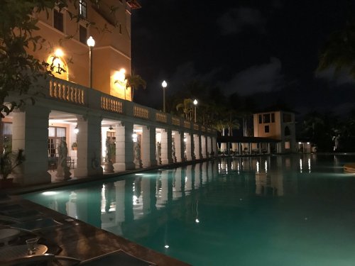 A serene nighttime view of a poolside area with illuminated buildings and palm trees reflecting in the water.