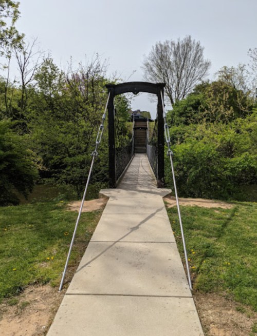 A suspension bridge leads into a lush green area, surrounded by trees and shrubs under a clear sky.