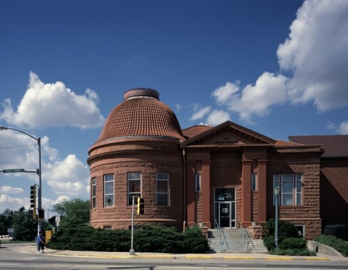 Historic brick building with a domed roof, surrounded by greenery and under a blue sky with fluffy clouds.