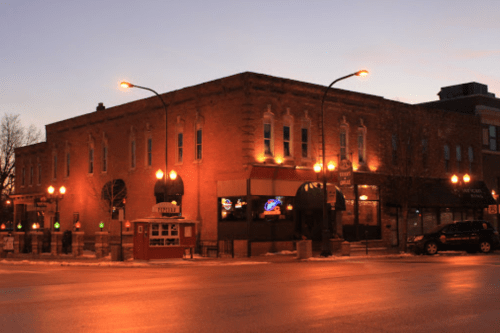 Historic brick building at dusk, featuring warm lights and a lively atmosphere, with parked cars and street lamps.