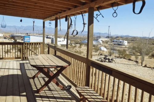 A wooden porch with a table, surrounded by desert scenery and hanging items, overlooking distant trailers and mountains.