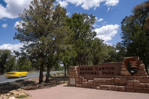 Sign for Grand Canyon National Park with trees and a blurred yellow car passing by on a sunny day.