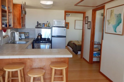 A cozy kitchen with wooden stools, a stove, and a fridge, leading to a small bedroom with a map on the wall.