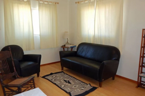 A cozy room with a black sofa, an armchair, a small table, and light curtains letting in natural light.