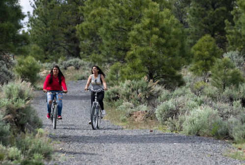 Two women ride bicycles on a gravel path surrounded by greenery and trees.