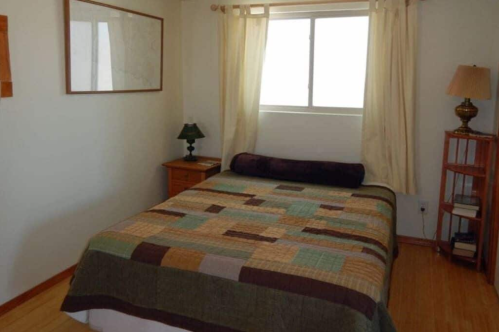 A cozy bedroom featuring a bed with a patterned quilt, a lamp, and a window with sheer curtains.