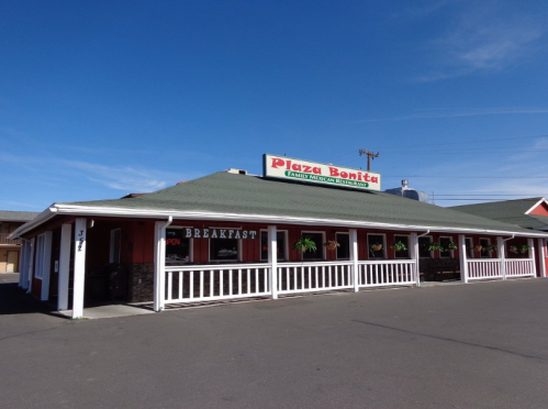 Exterior of Plaza Bonita restaurant with a green roof, white railings, and a sign advertising breakfast. Clear blue sky.