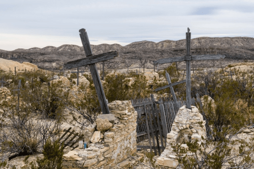 Abandoned graveyard with wooden crosses and stone ruins, surrounded by desert landscape and distant mountains.