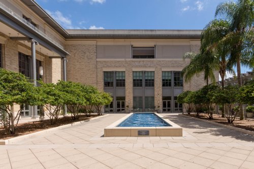A courtyard with a small water feature, surrounded by greenery and a modern building under a clear blue sky.