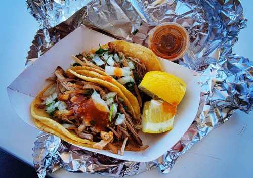 A takeout box with three tacos filled with meat, onions, and cilantro, accompanied by a lemon wedge and sauce.