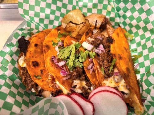 A close-up of three tacos with meat, cheese, and toppings, served in a green checkered basket with radishes on the side.