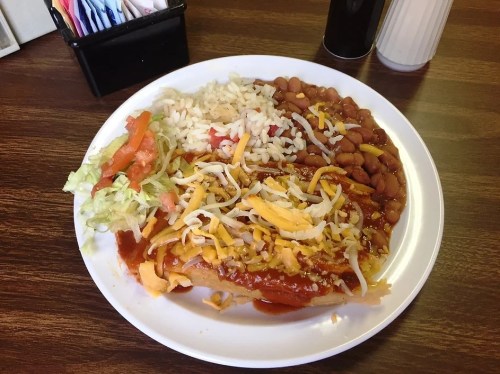 A plate of enchiladas topped with cheese, served with rice, beans, and a side of lettuce and tomato.
