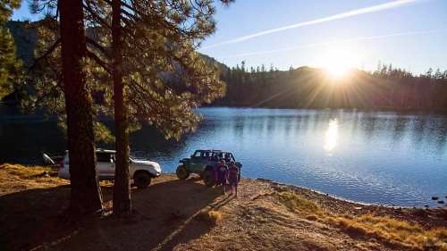 Two vehicles parked by a serene lake at sunset, surrounded by trees and mountains in the background.