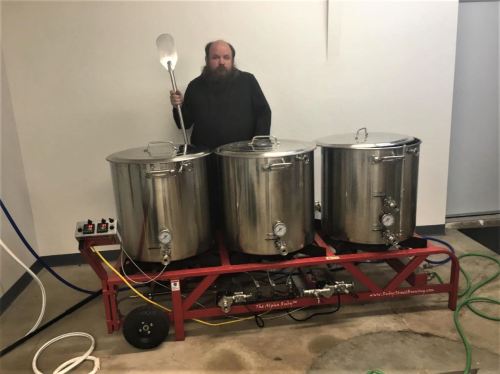 A person stands beside a three-vessel brewing system on wheels, holding a large spoon, in a workshop setting.