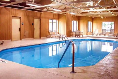 Indoor swimming pool with lounge chairs, wooden walls, and large windows letting in natural light.