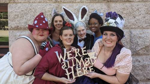 A group of seven people in whimsical costumes poses together, holding a sign that says "We're all mad here."