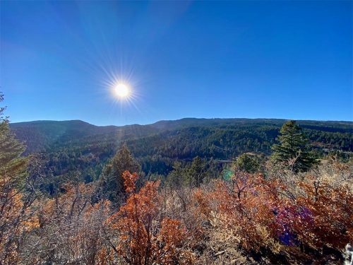 A sunlit landscape with rolling hills, dense forests, and colorful autumn foliage under a clear blue sky.