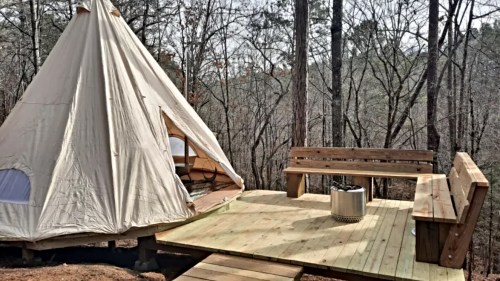 A canvas tent on a wooden deck surrounded by trees, featuring a fire pit and a bench for seating.