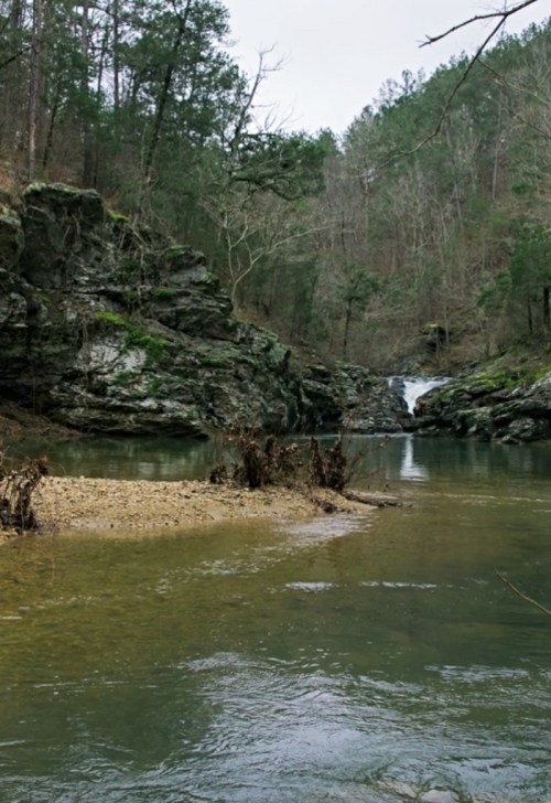 A serene river scene with rocky banks, trees, and a small waterfall in the background.