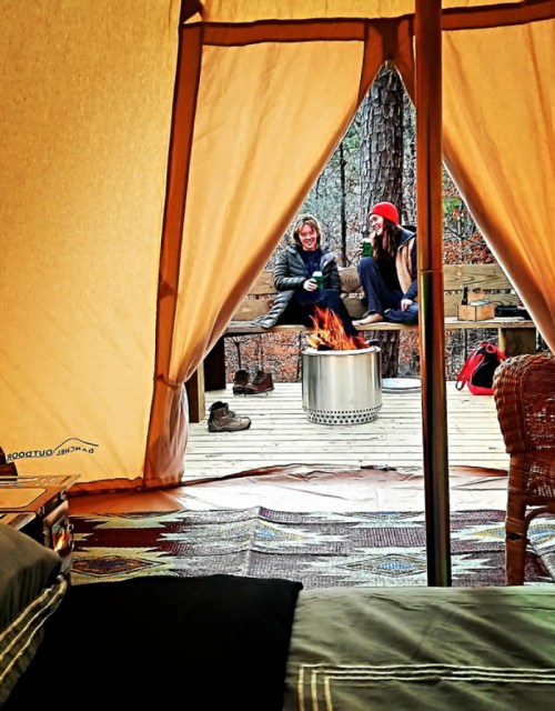 Two women sit by a fire outside a tent, enjoying drinks and conversation in a cozy, wooded campsite.