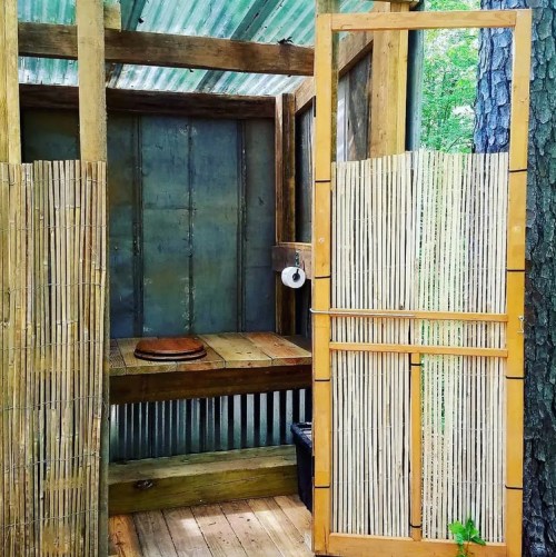A rustic outdoor bathroom with wooden walls, a bamboo screen, and a wooden bench, surrounded by trees.