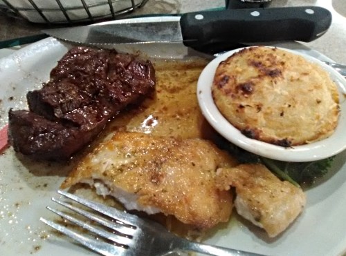 A plate with a piece of steak, fried fish, and a small dish of baked potato, accompanied by a knife and fork.