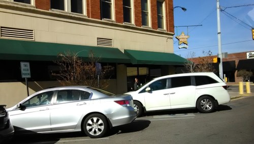 A silver car and a white minivan parked on a street in front of a building with green awnings.