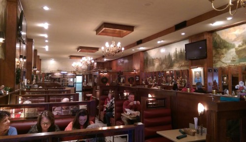 A cozy diner interior with wooden booths, chandeliers, and muraled walls, filled with patrons enjoying their meals.
