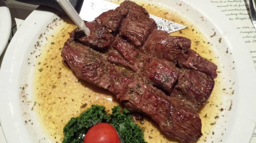 A plate of grilled steak slices garnished with herbs, served with a cherry tomato and leafy greens.