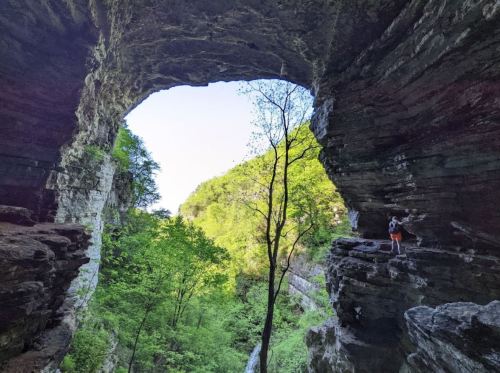 A natural rock arch frames a lush green landscape with a person standing on a ledge, surrounded by trees.