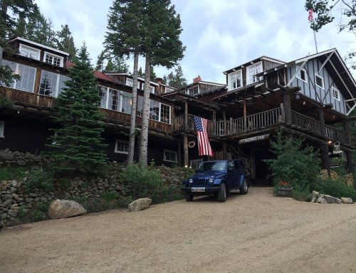 A rustic lodge with wooden architecture, surrounded by trees, featuring an American flag and a blue Jeep parked in front.
