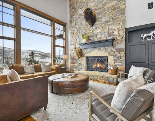 Cozy living room with stone fireplace, large windows, and mountain views, featuring comfortable seating and rustic decor.