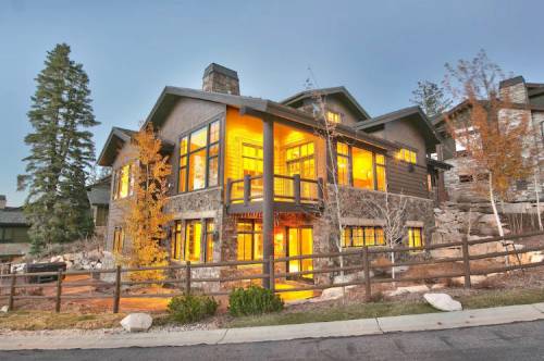 A modern mountain home with large windows glowing warmly, surrounded by trees and a stone pathway.