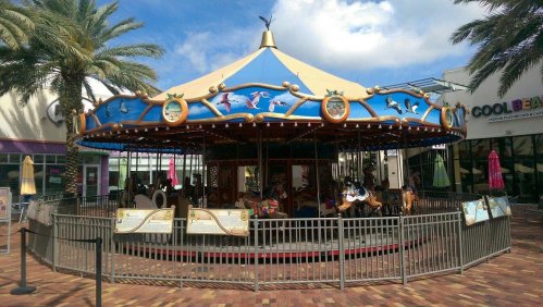 A colorful carousel with ornate decorations, surrounded by palm trees and a shopping area in the background.