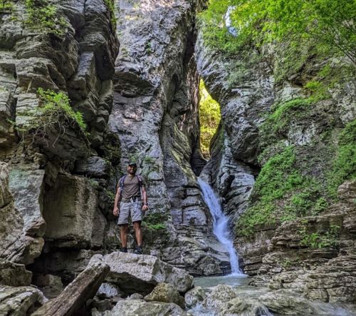 A person stands on a rock in a narrow canyon with a waterfall and lush greenery surrounding the area.