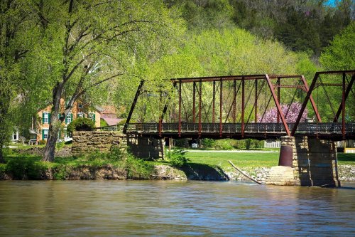 A rusty iron bridge spans a river, surrounded by lush green trees and a quaint house in the background.