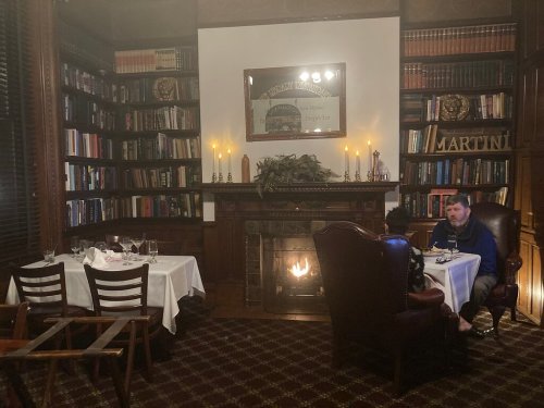 Cozy restaurant interior with bookshelves, a fireplace, and two tables set for dining. Warm, inviting atmosphere.