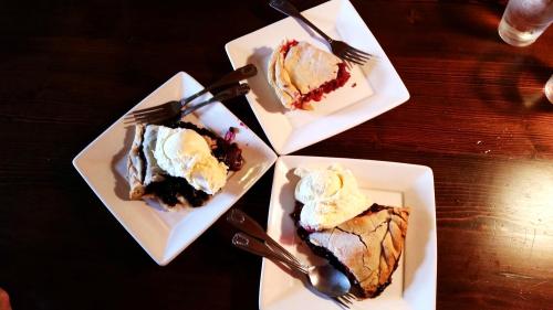 Three slices of pie topped with ice cream on white plates, arranged on a wooden table. Forks and spoons are beside each slice.