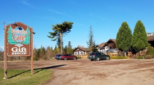 A rustic café and gift shop with a sign, surrounded by trees and parked cars on a sunny day.