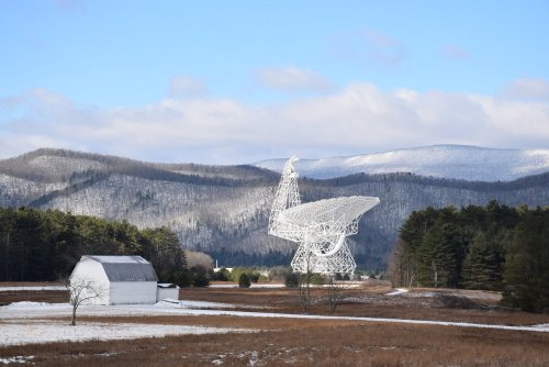 A large white radio telescope stands beside a barn in a snowy landscape, with mountains in the background.
