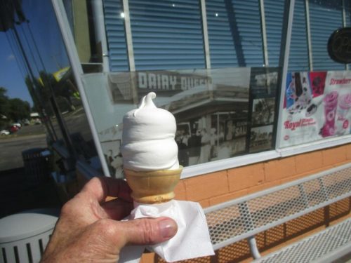 A hand holding a soft serve ice cream cone in front of a Dairy Queen with vintage photos on the wall.