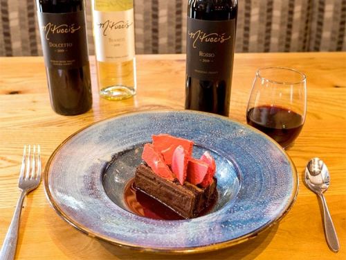 A slice of chocolate cake with raspberry garnish on a decorative plate, accompanied by wine bottles and a glass of red wine.
