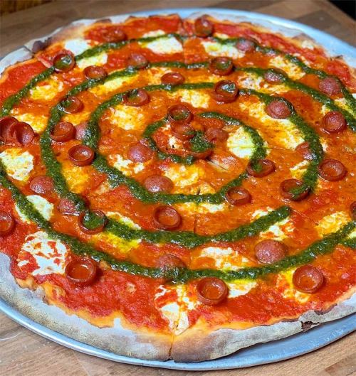 A circular pizza topped with spiral patterns of tomato sauce, cheese, pepperoni, and green pesto.